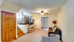 Un vaste espace de vie doté d’une porte en bois, d’un escalier, d’un ventilateur de plafond, d’un téléviseur posé sur un meuble, d’une console blanche, d’un fauteuil gris et d’un canapé.