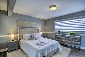 A modern bedroom with a tufted headboard, a large bed with white bedding, a nightstand with a lamp, a dresser with drawers, and a window with blinds. - Cozy Port Orchard Home w/ Grill & Fire Pit! (Port Orchard)