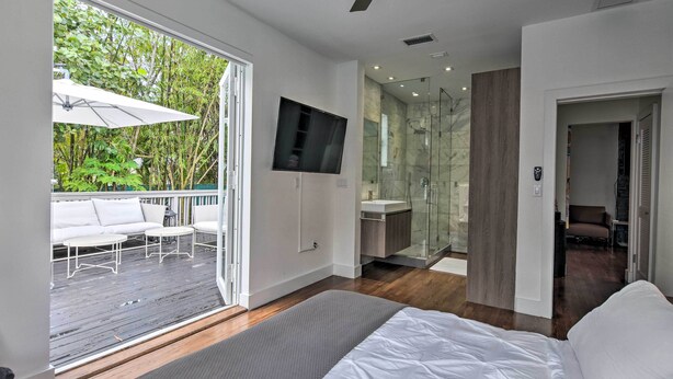 A modern bedroom with a large bed, a TV mounted on the wall, a glass shower, and a balcony with outdoor seating. - Modern Miami Villa w/ Pool Oasis ~ 5 Mi to Beach! (North Miami)