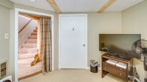 Une pièce avec une porte blanche, un escalier orné d’un rideau à carreaux, un téléviseur à écran plat, un ventilateur sur pied et une petite poubelle.