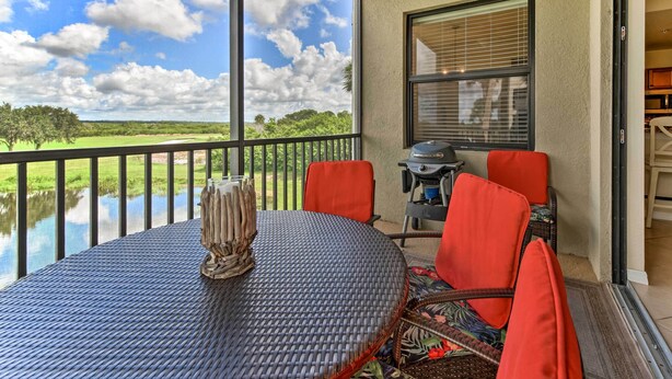 A balcony with a round table, red chairs, and a view of a lake and trees. - Screened Patio & Resort Perks: Bradenton Condo! (Bradenton)