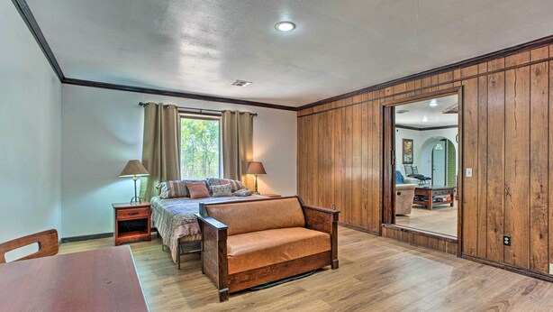 A room with a wooden floor, a sofa, a small table, and a bed. There is a window with curtains and a lamp on a nightstand. - Lovely 'anna Fink House' w/ Patio & Fire Pit (Atoka)