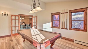 A pool table in a room with wooden floors, a kitchen area visible through a glass door, and a large window with curtains. - Mtn View + Group Ready: Conifer Home on 100 Acres! (Conifer)