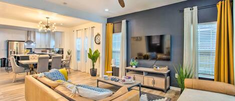 A modern living room with a sofa, coffee table, and TV. There is a dining area with chairs and a table, and a kitchen in the background.