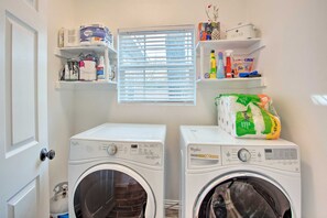 Une buanderie équipée de deux lave-linge Whirlpool, d’étagères contenant des produits de nettoyage et d’une fenêtre avec des stores.