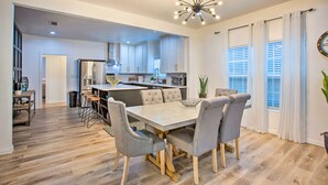 Una cocina moderna con armarios blancos, una isla central y un comedor con una mesa de tonos claros y sillas grises.