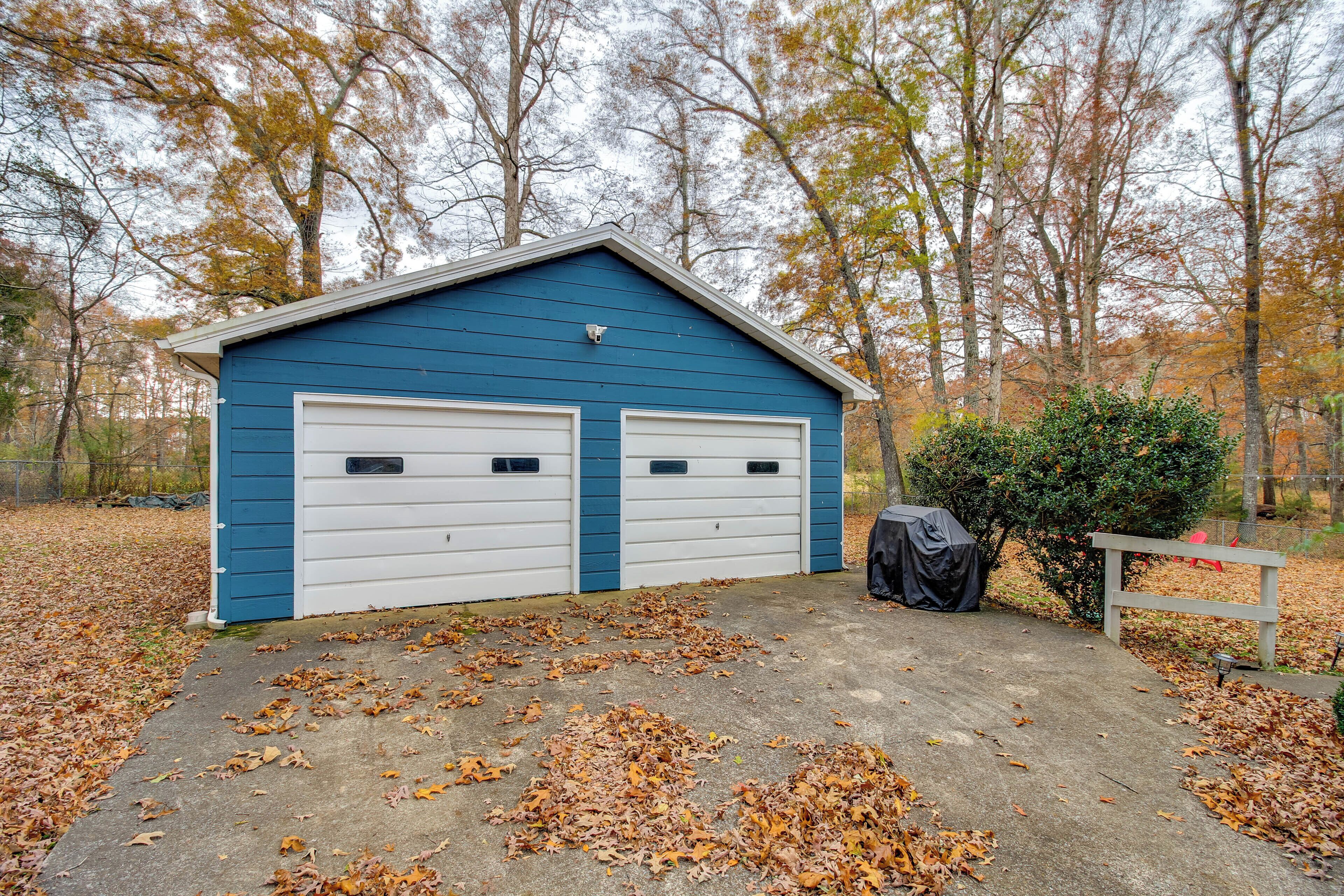 A blue garage with two white doors, surrounded by fallen leaves and trees.