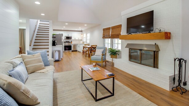 Una sala de estar moderna con un sofá, una mesa de centro y una chimenea. Se observa una cocina al fondo, con una zona de comedor y una escalera que sube a otro nivel.
