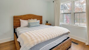 Un dormitorio con cabecera de mimbre, cama con manta a rayas, almohada con texto, una mesita con planta y ventana con cortinas.