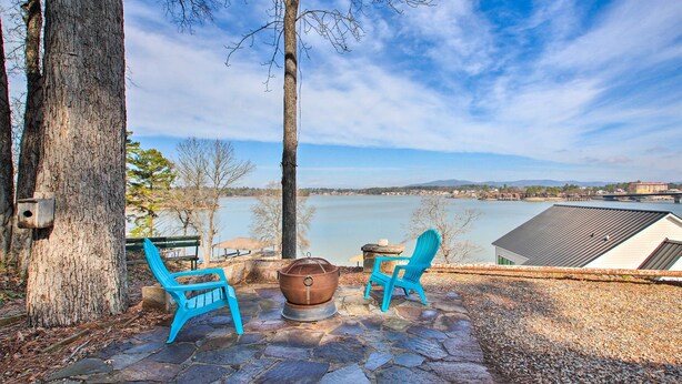 A stone patio with two blue Adirondack chairs and a fire pit, overlooking a lake and distant buildings.