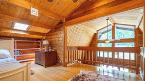 Ein Schlafzimmer in einer Blockhütte mit einem Bett, einer Kommode, einem Kuhfellteppich und einem großen Fenster mit Blick auf Bäume.