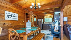 Ein Holzblockhaus-Interieur mit Esstisch, Stühlen, einer Bank, einer Küchenzeile und einem Schild mit der Aufschrift 'Kommt und versammelt euch an unserem Tisch'.