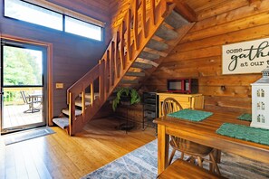 A wooden interior with a staircase, a dining table with chairs, a sign saying "come gather at our table," and a view of the outdoors through a door. - Rustic Fancy Gap Vacation Rental With Fire Pit (Fancy Gap)