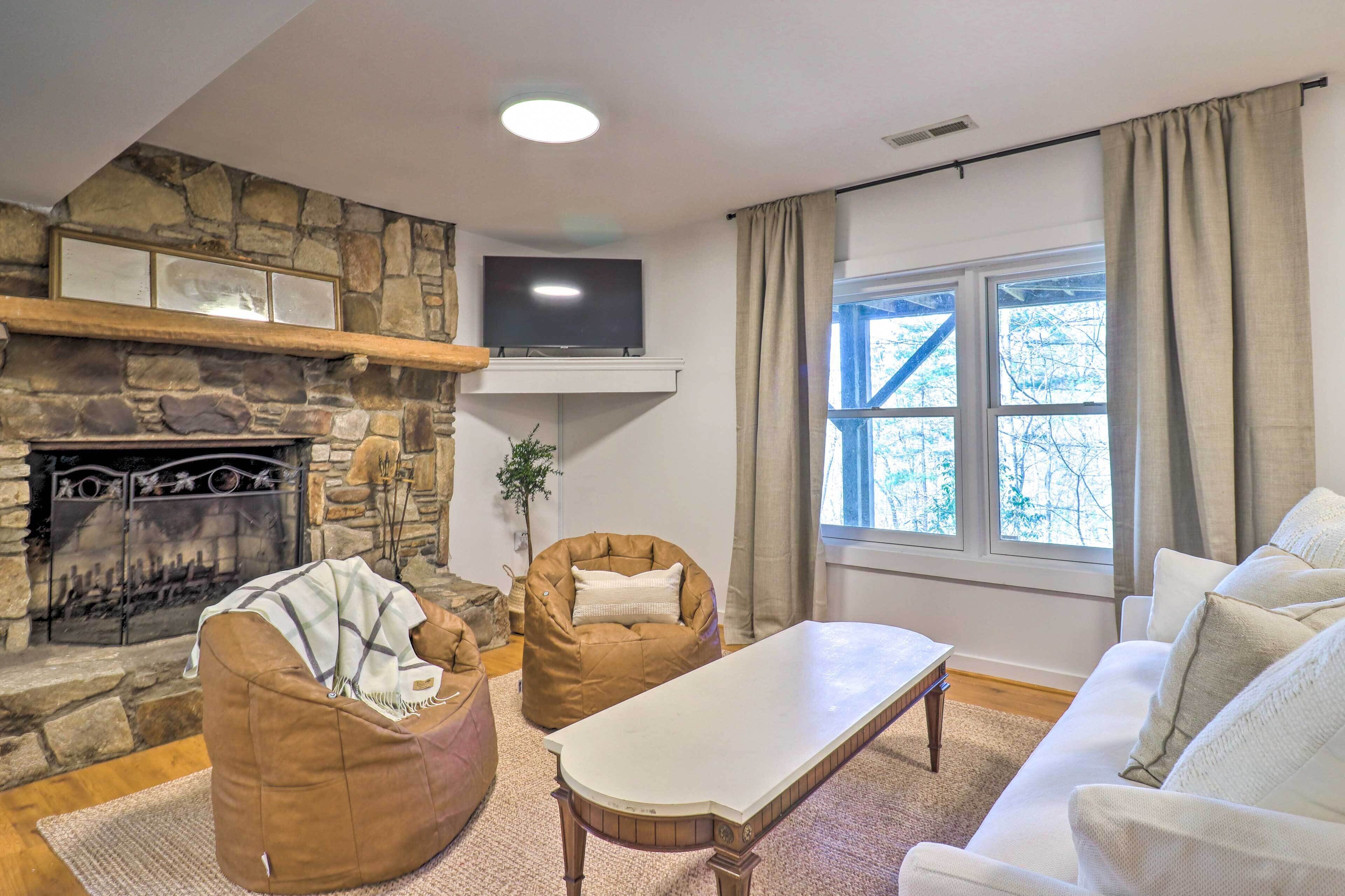 A cozy living room with a stone fireplace, a flat-screen TV, comfortable seating, and a large window with curtains.