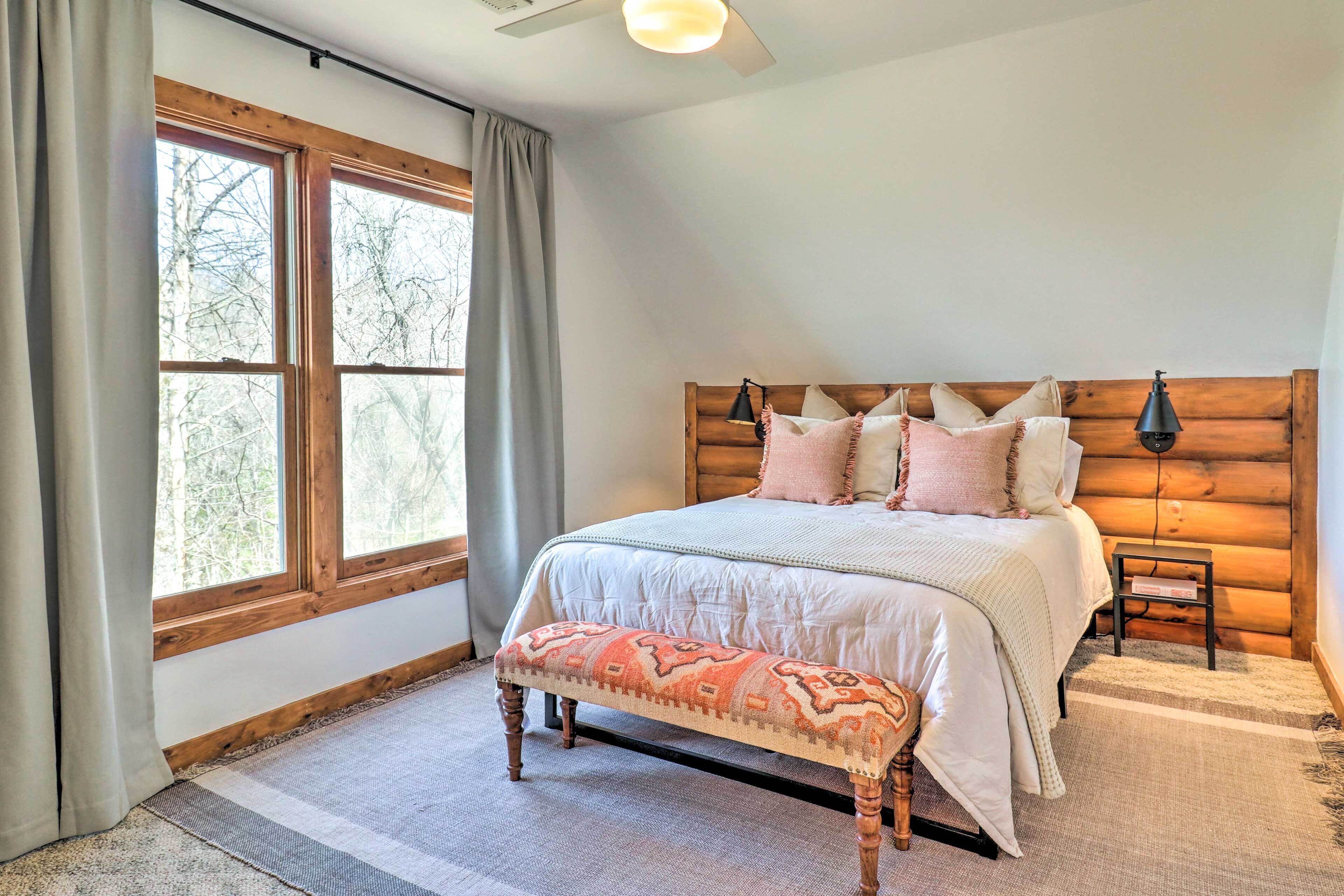 Une chambre à coucher avec un grand lit, une tête de lit en bois, un banc, une table de chevet et une fenêtre avec des rideaux.