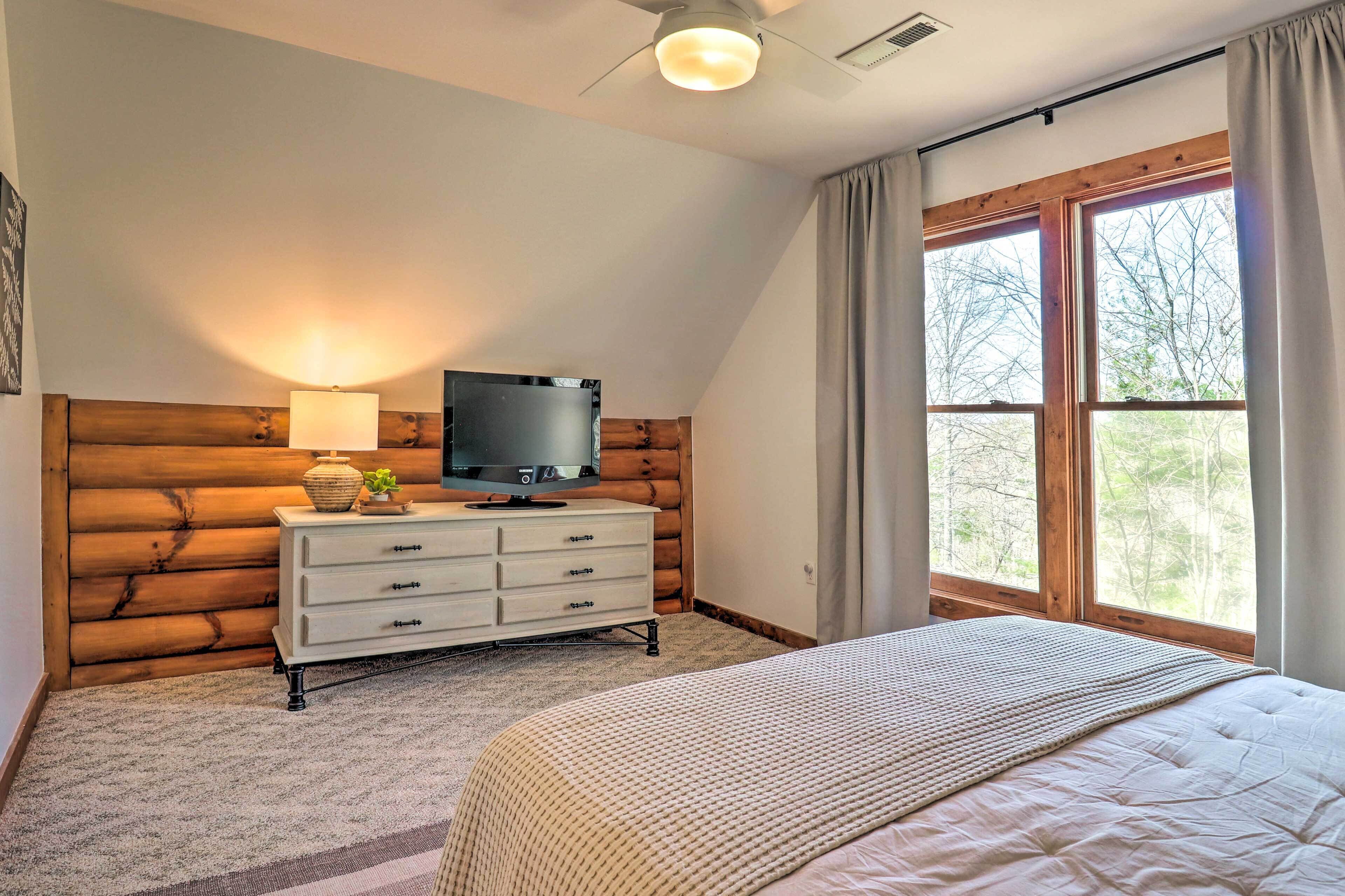 Une chambre à coucher avec une tête de lit en bois, un téléviseur posé sur une commode, une grande fenêtre avec des rideaux et un lit recouvert d’une couette blanche.