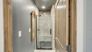 Un pasillo estrecho con ducha envuelta en cristal, puertas de madera y un espejo en la pared izquierda.