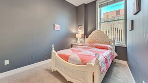 A bedroom with a bed, a nightstand, a lamp, and a window with a view of buildings. - Heart of Dtwn Omaha: Historical Apt w/ Perks (Omaha)