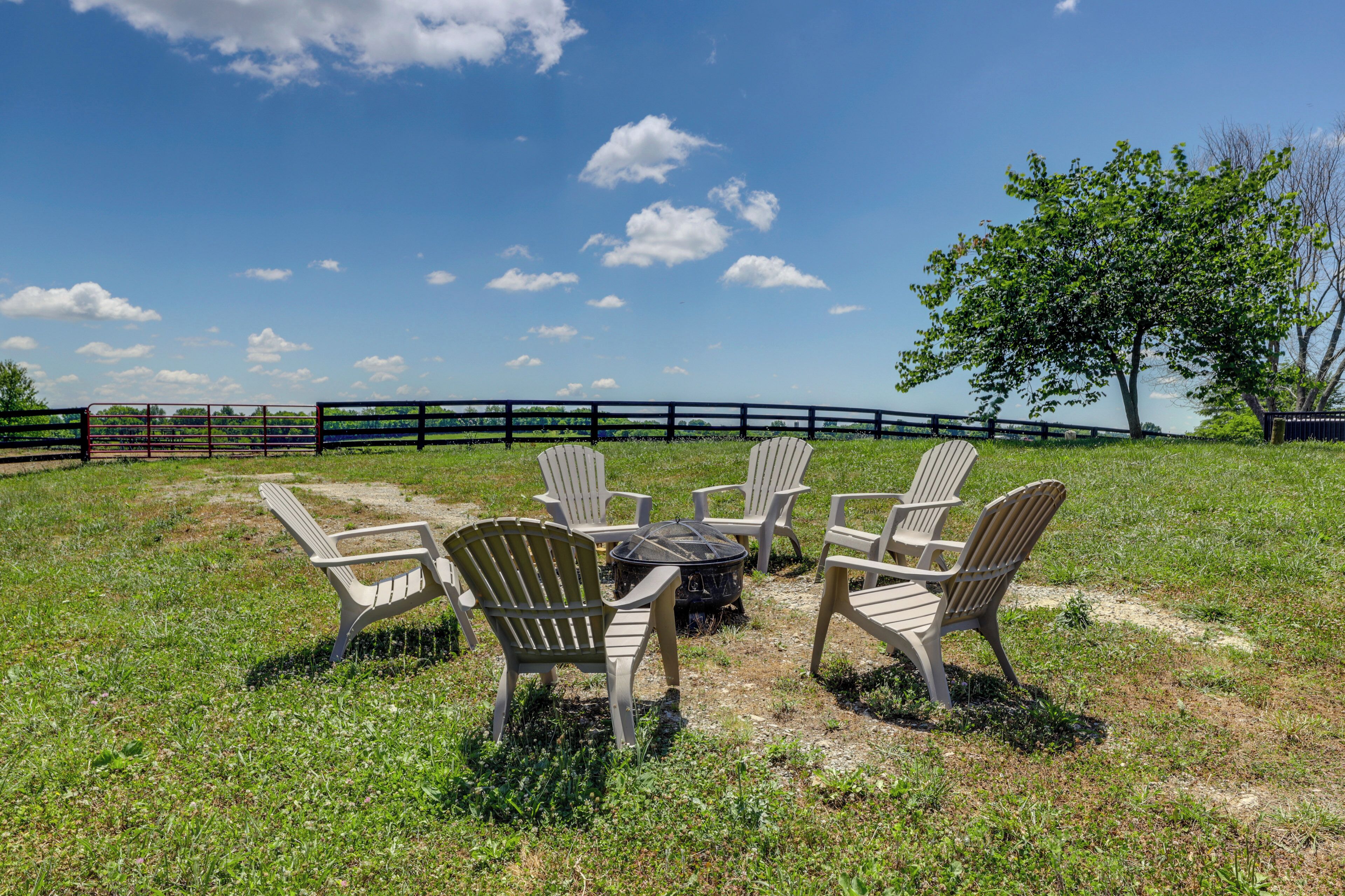 An outdoor seating area with Adirondack chairs and a fire pit, surrounded by a grassy field and a fence.