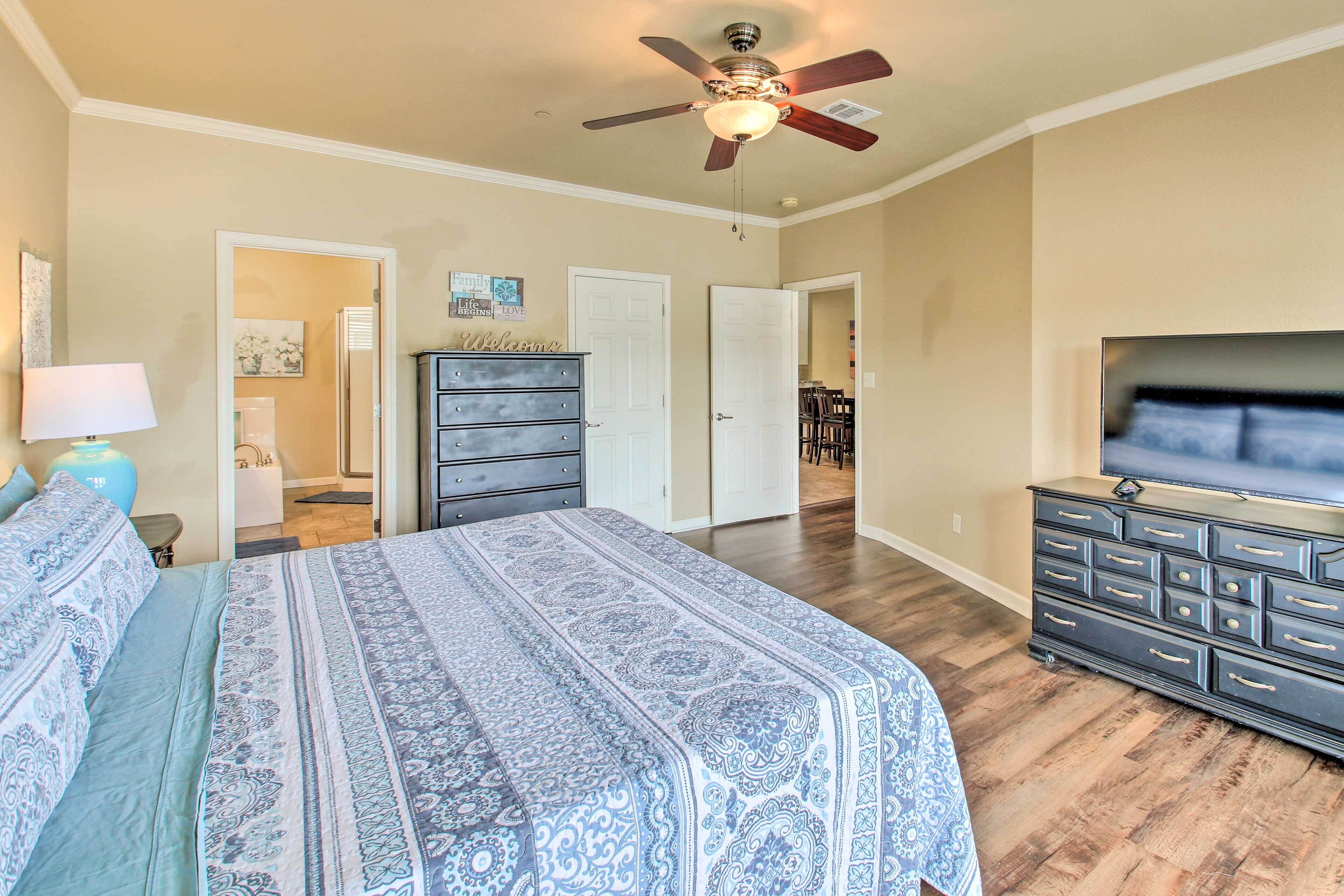 A bedroom with a bed, dresser, TV, ceiling fan, and a hallway leading to a bathroom.