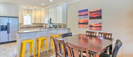 A modern kitchen with white cabinets, a stainless steel refrigerator, and a dining area with a wooden table and yellow stools.
