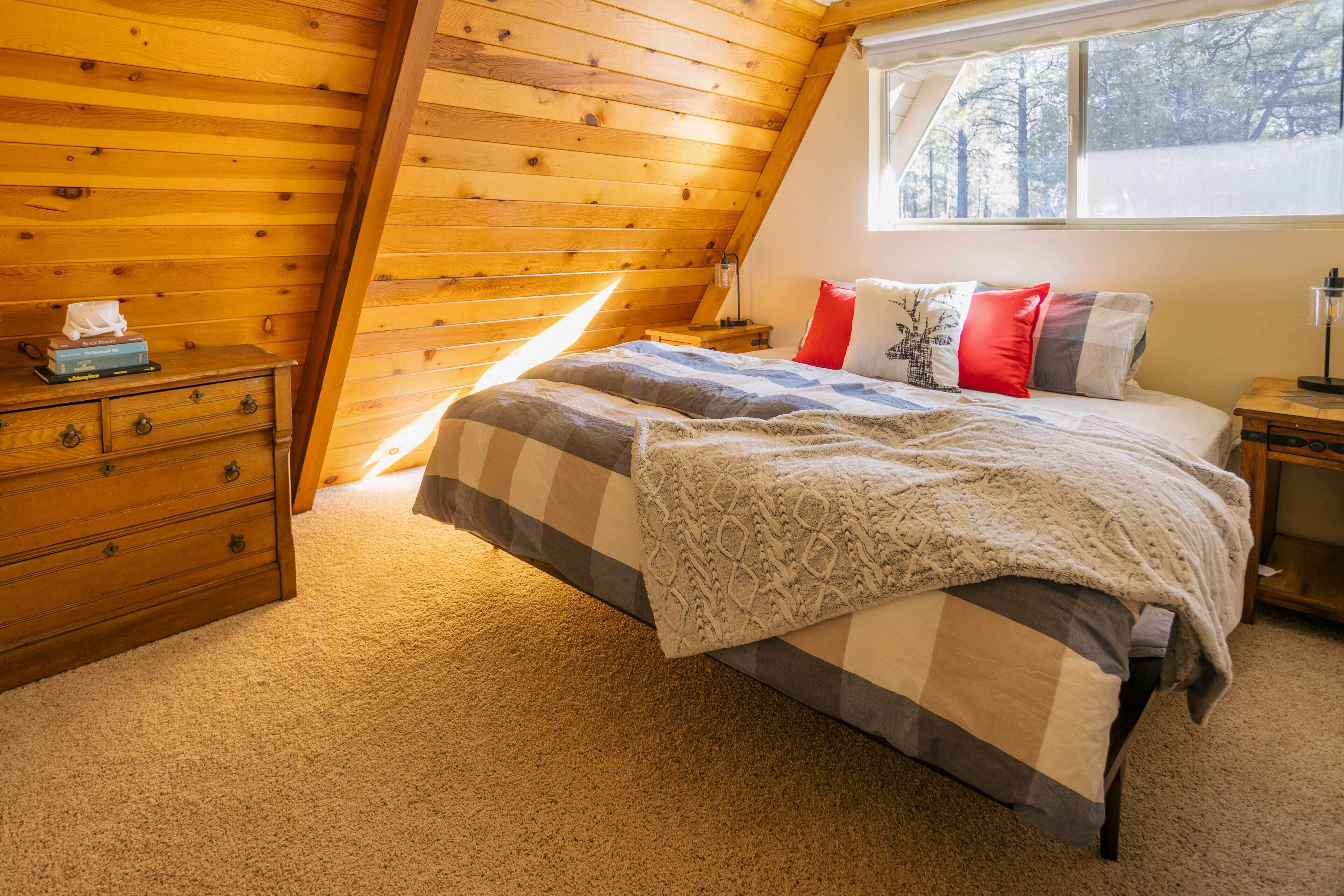 A cozy bedroom with a sloped ceiling, a bed with multiple pillows, a wooden dresser, and a window with a view of trees.