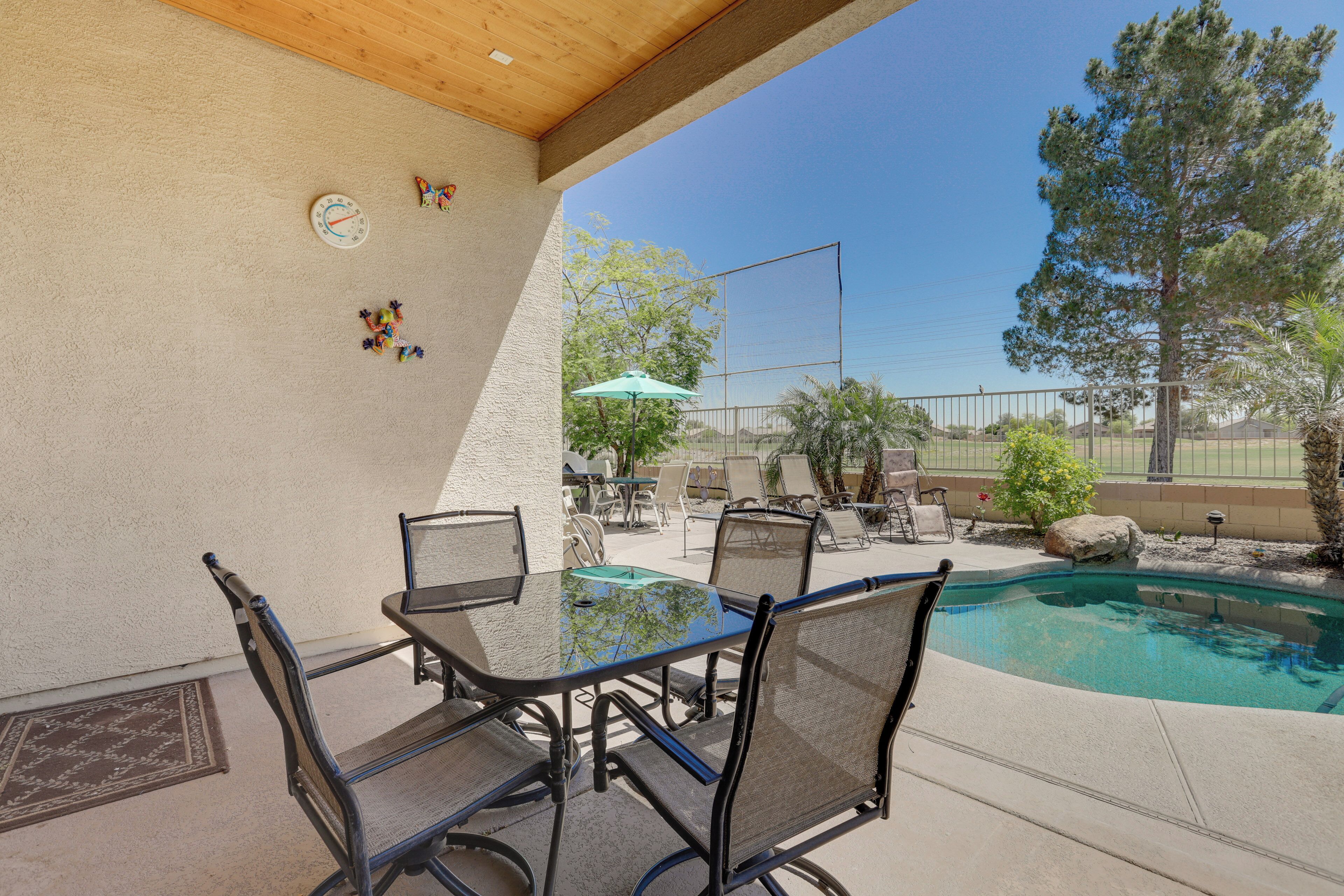 Un patio exterior con mesa y sillas de vidrio, una piscina y una cancha de tenis al fondo.