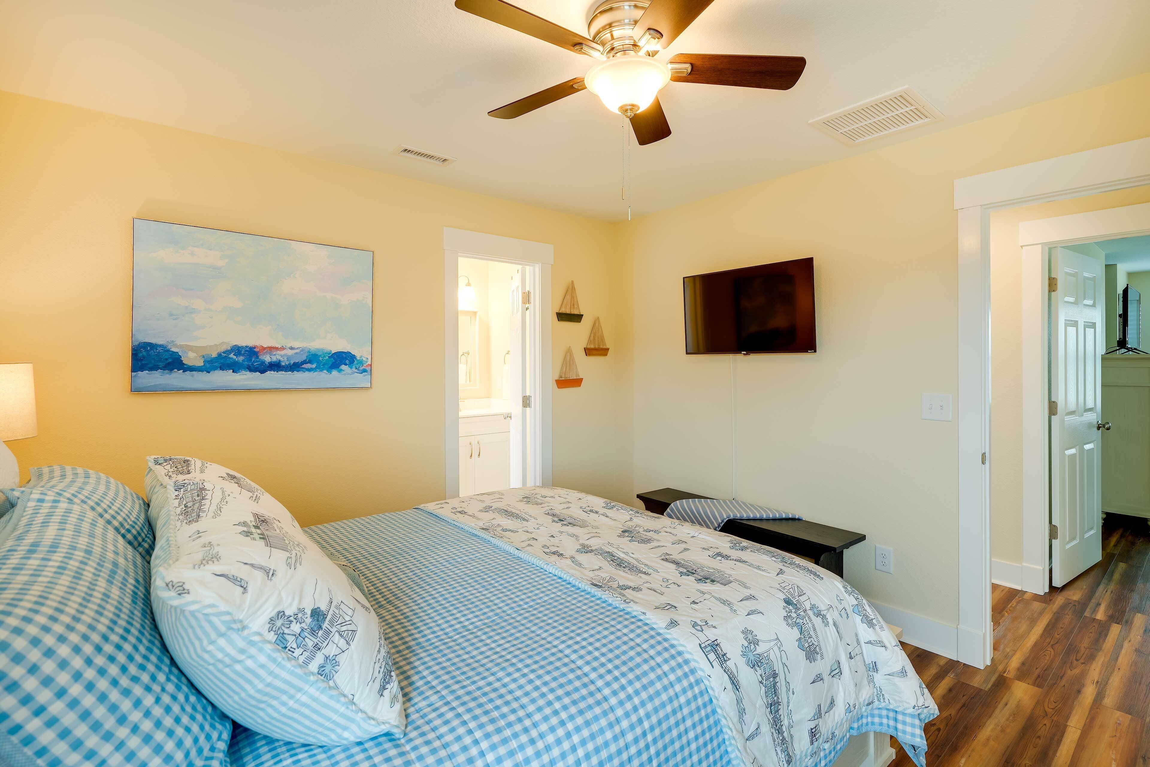 A bedroom with a bed, a ceiling fan, a painting, a television, and a door leading to another room.