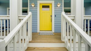 A wooden staircase leading to a bright yellow door with a small window, flanked by white railings and two wall-mounted lights. - Corolla Vacation Rental w/ Pool, Walk to Beach! (Corolla)