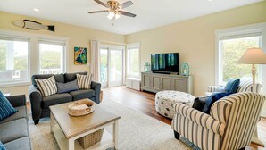 A spacious living room with a ceiling fan, a television on a cabinet, and a large window. - Corolla Vacation Rental w/ Pool, Walk to Beach! (Corolla)
