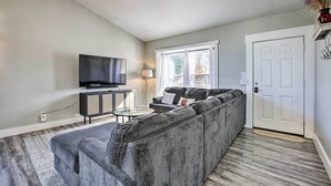 Ein Wohnzimmer mit einem grauen Ecksofa, einem Flachbildfernseher, einem kleinen runden Couchtisch und einer Lampe.