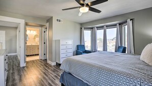 Ein Schlafzimmer mit einem Bett, einem Deckenventilator, einem Fenster mit Vorhängen, einem Sessel und Blick ins Freie.
