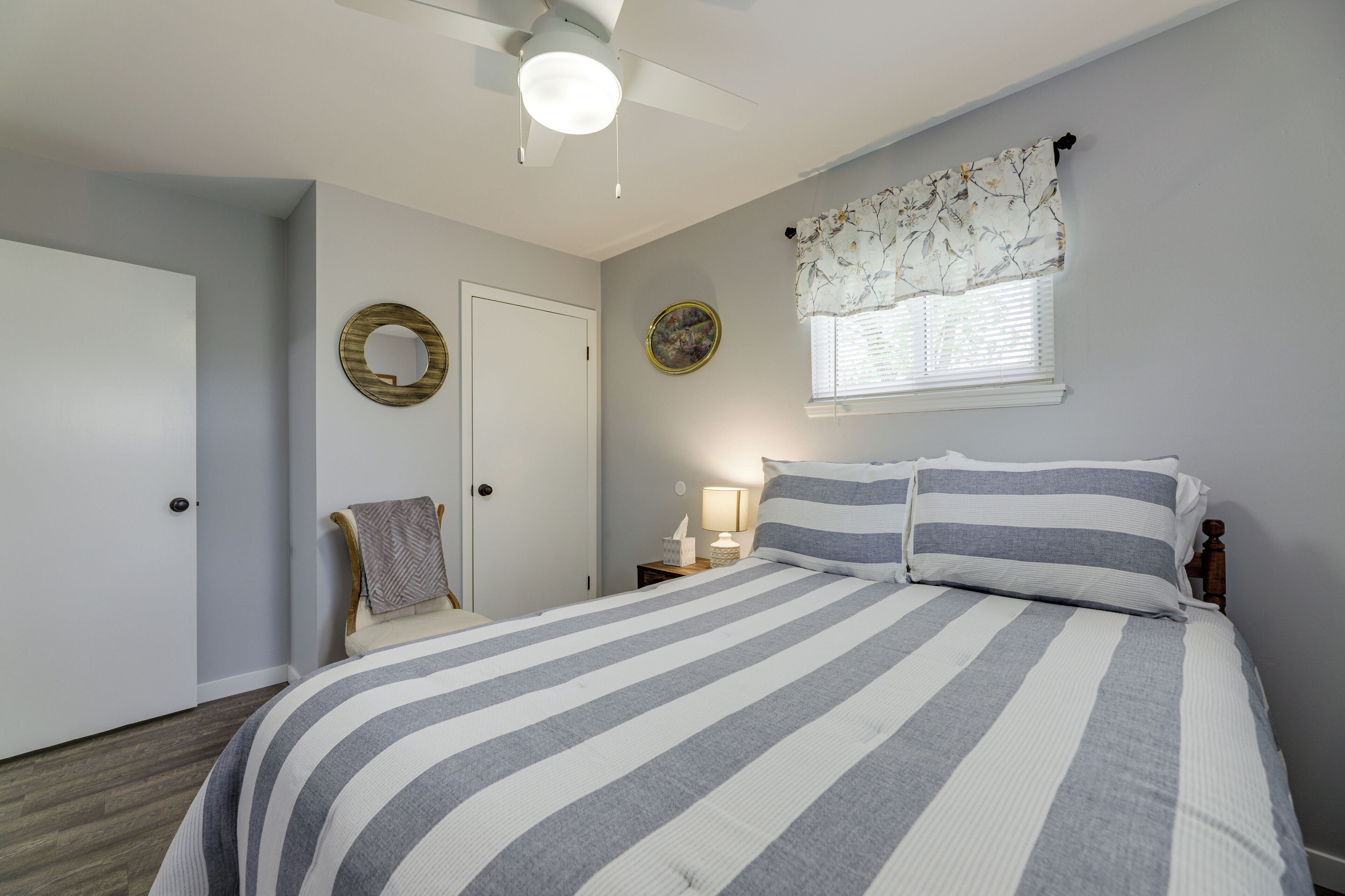 Une chambre à coucher comprenant un lit, une table de chevet, une chaise, une porte, une fenêtre avec des rideaux et un ventilateur de plafond.