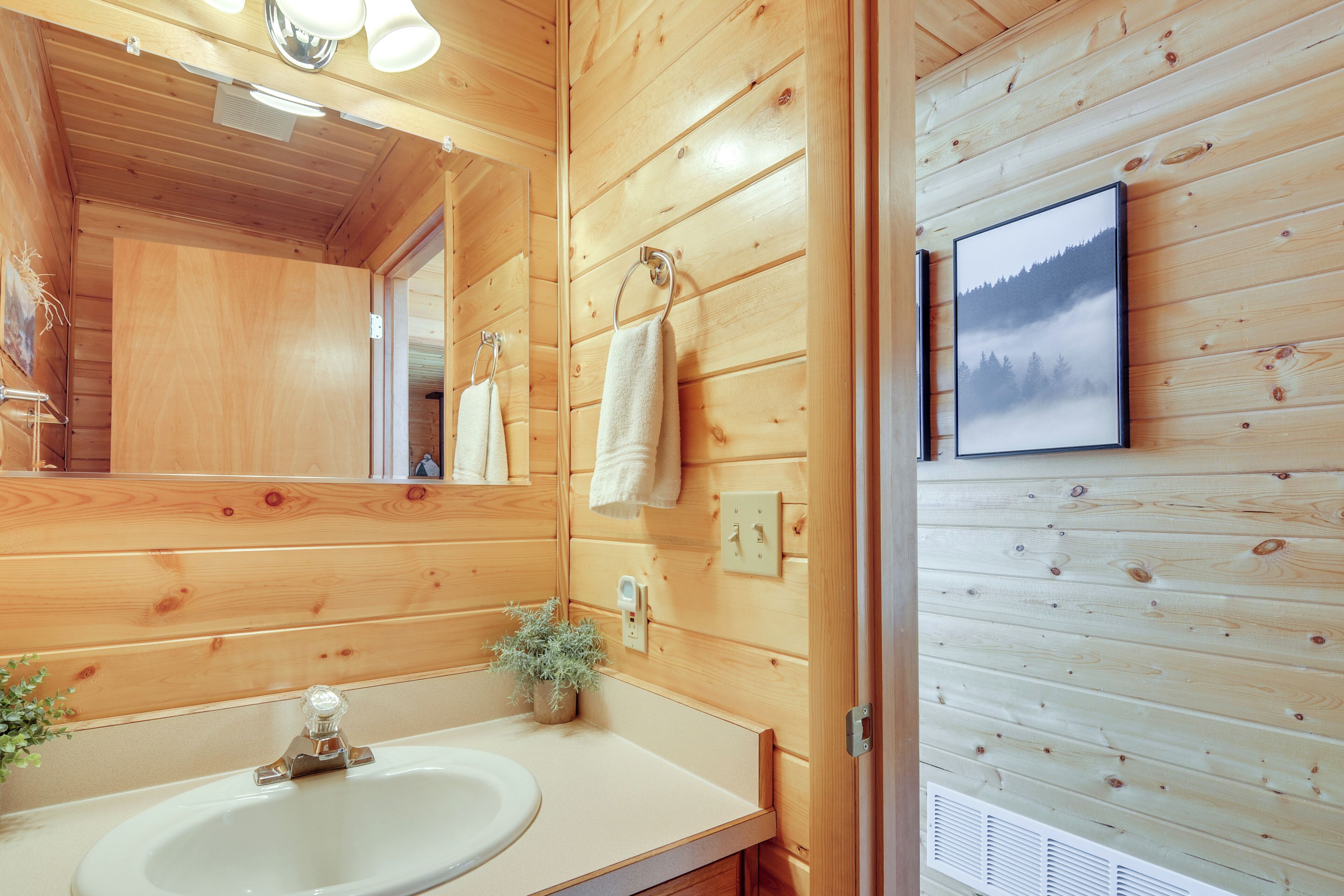 Une salle de bain rustique, aménagée dans une cabane en bois, avec un lavabo, un miroir, un porte-serviettes et un tableau encadré accroché au mur.