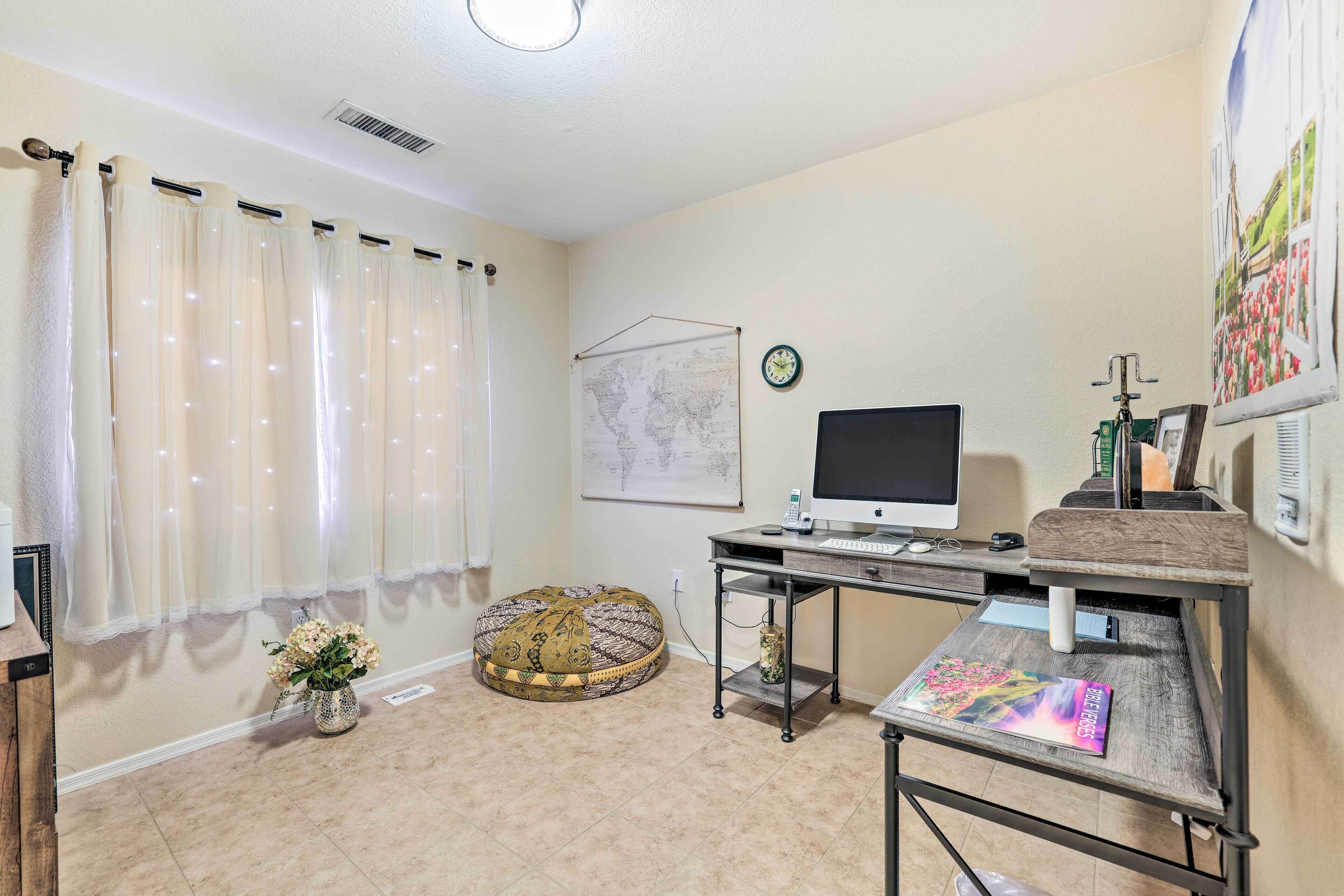 Una habitación con un escritorio, computadora y un mapa en la pared. Hay una ventana con cortinas, un otomanillo redondo y un jarrón con flores.