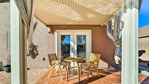 Eine Terrasse im Freien mit einem Holztisch und Stühlen, einem überdachten Bereich und einer Tür zu einem Hauseingang.