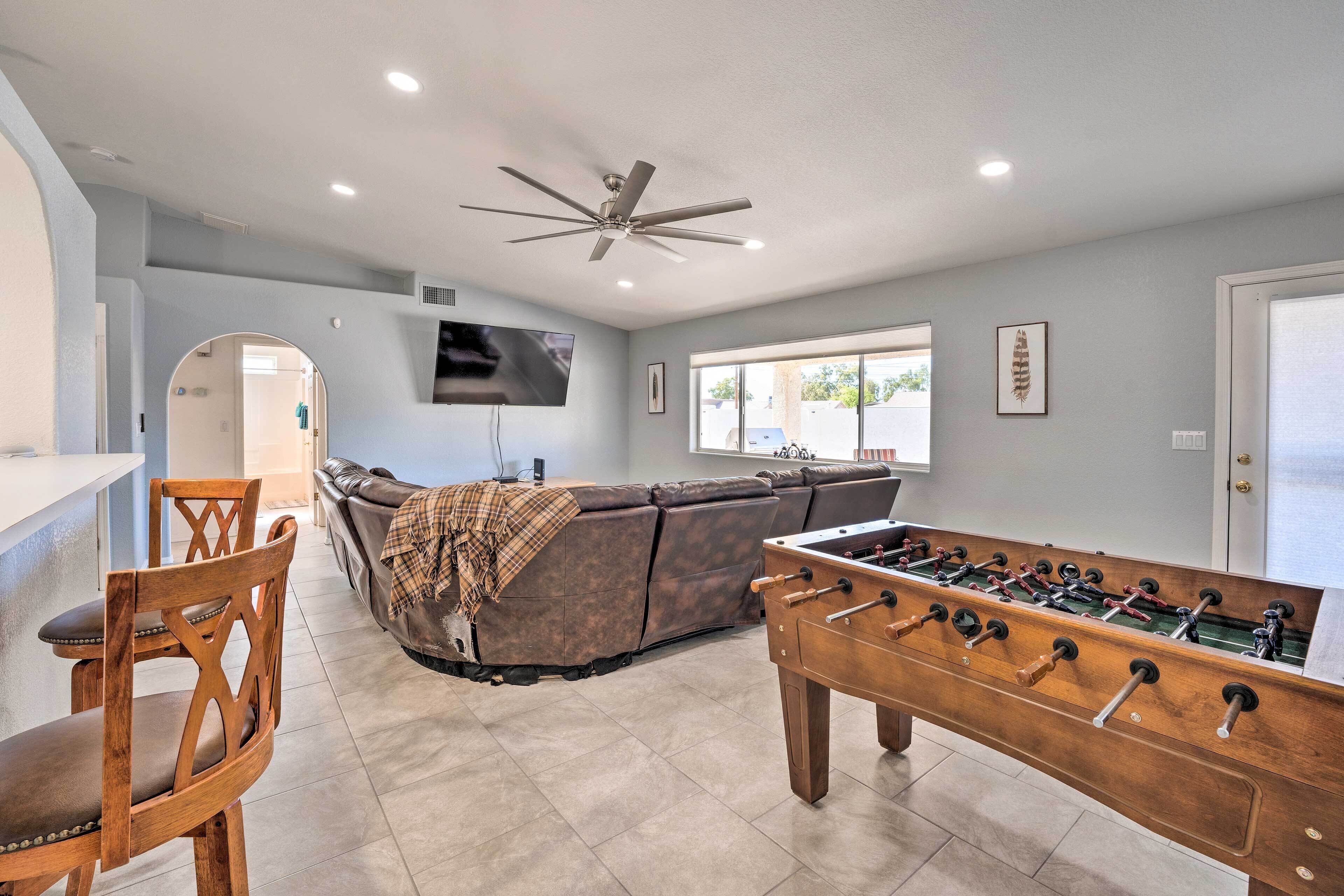 A modern living room with a foosball table, a sofa, a TV mounted on the wall, and a ceiling fan.