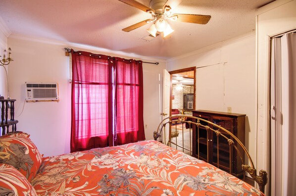 Un dormitorio con cama, ventilador de techo, ventana con cortinas rojas, cabecero de madera y un espejo en la pared.