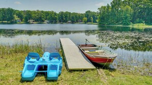 Un molo con una piccola imbarcazione e una boa gonfiabile blu, accanto a un lago tranquillo circondato da alberi e case.