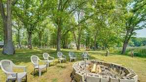 Boccia in cemento per il fuoco, circondata da sedie bianche in plastica, posta in una zona erbosa con alberi.