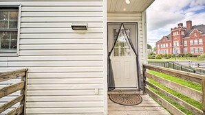 Een wit huis met een houten veranda, een witte deur met een zwart gordijn, en een rode bakstenen gebouw op de achtergrond.