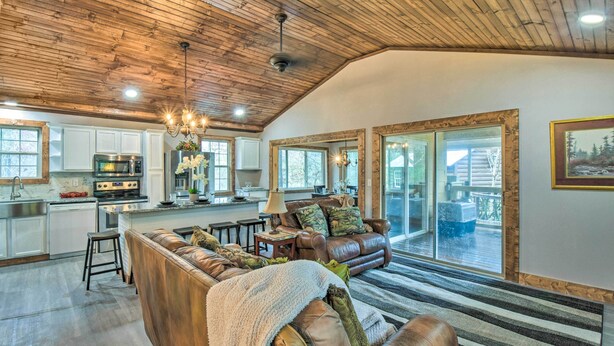 A spacious living room with a leather sofa, a coffee table, and a chandelier. There is a kitchen area with modern appliances and a large window. - Otto Home w/ Fire Pit, Tree House & Hot Tub! (Otto)