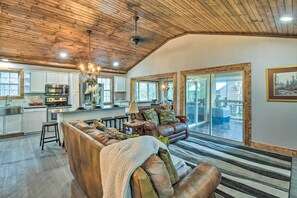 A spacious living room with a leather sofa, a coffee table, and a chandelier. There is a kitchen area with modern appliances and a large window. - Otto Home w/ Fire Pit, Tree House & Hot Tub! (Otto)