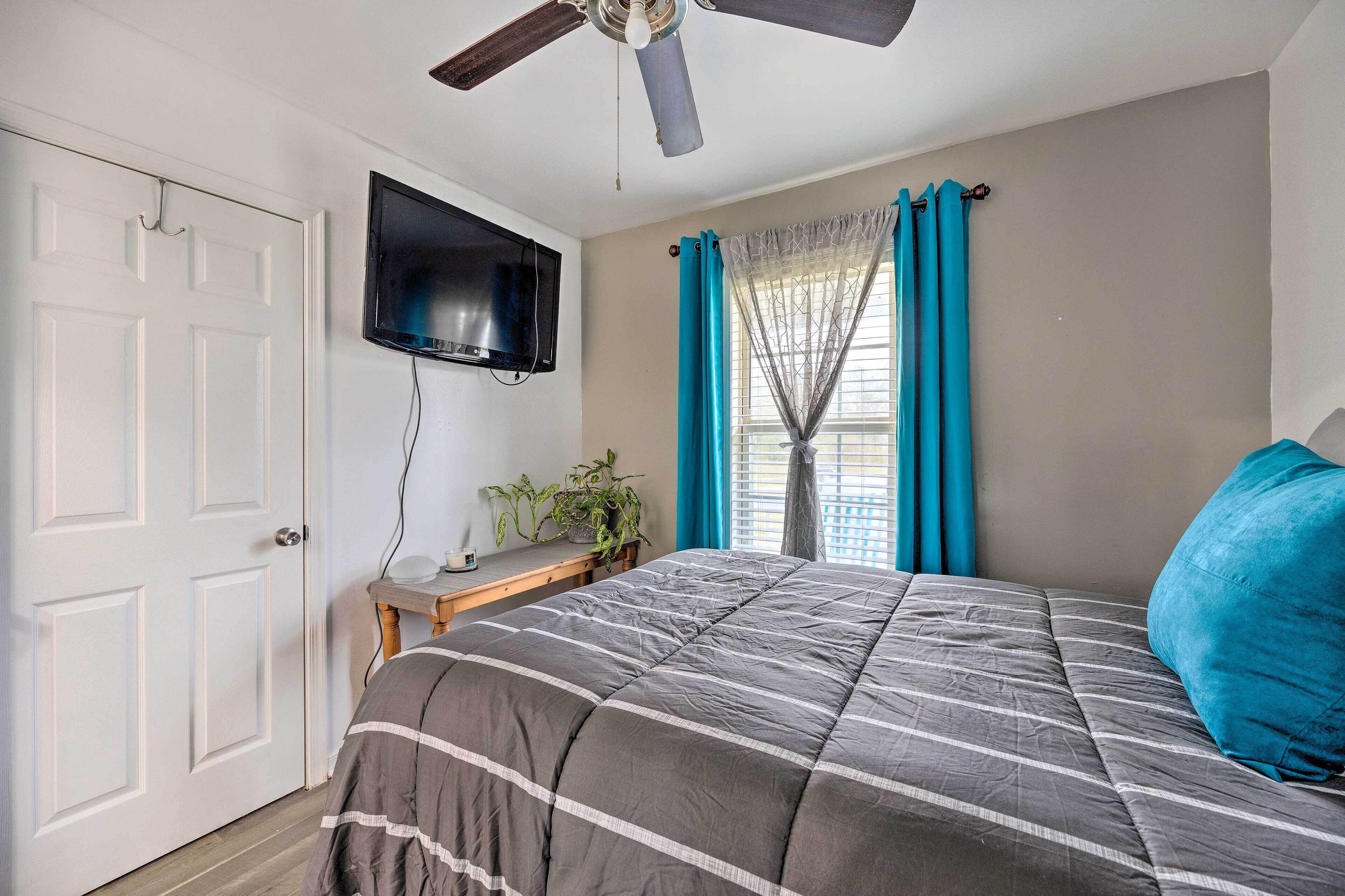 Ein Schlafzimmer mit einem Bett, einem Deckenventilator, einem an der Wand montierten Fernseher und einem Fenster mit Vorhängen.
