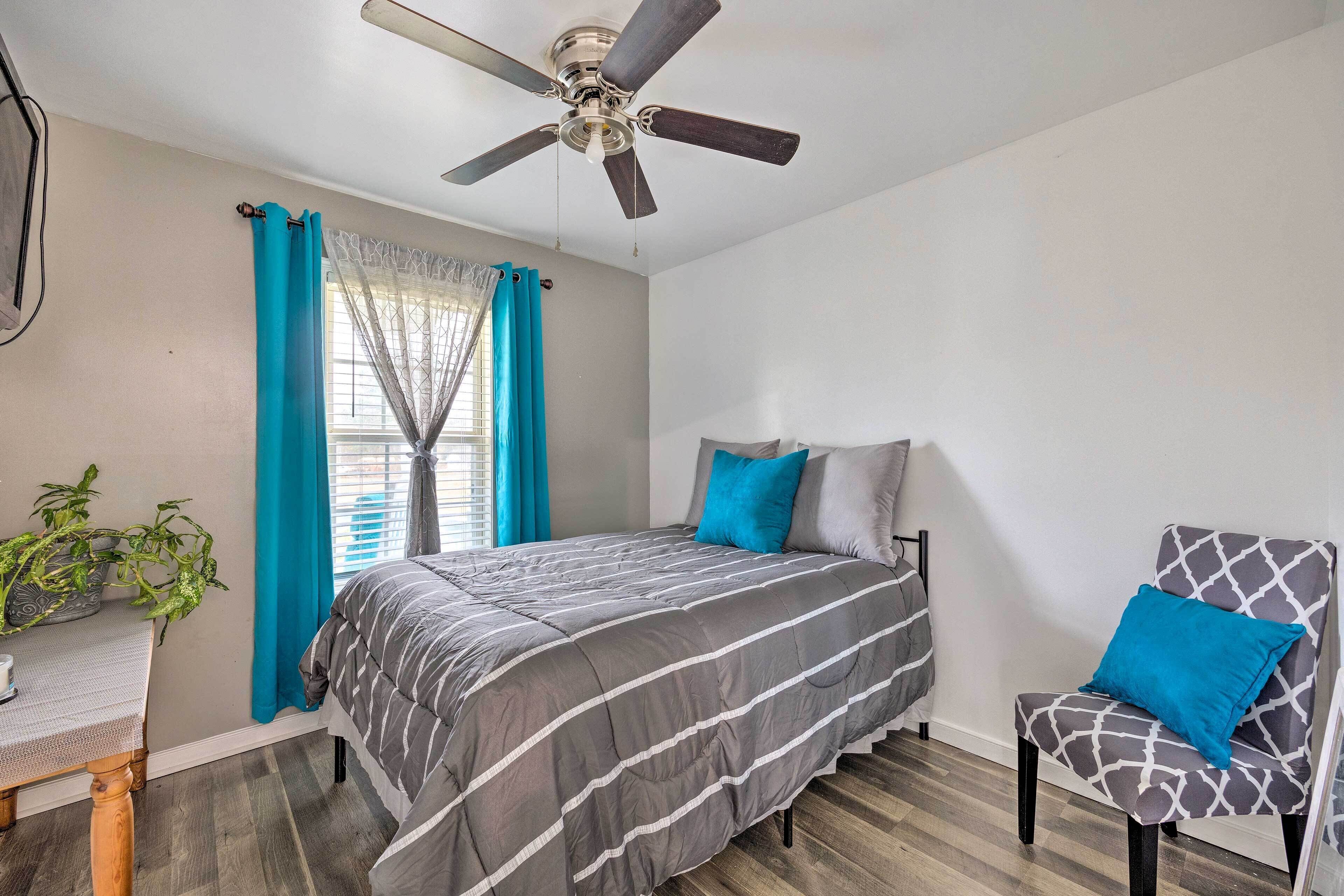 Un dormitorio con cama, una silla, ventilador de techo, ventana con cortinas y una planta.
