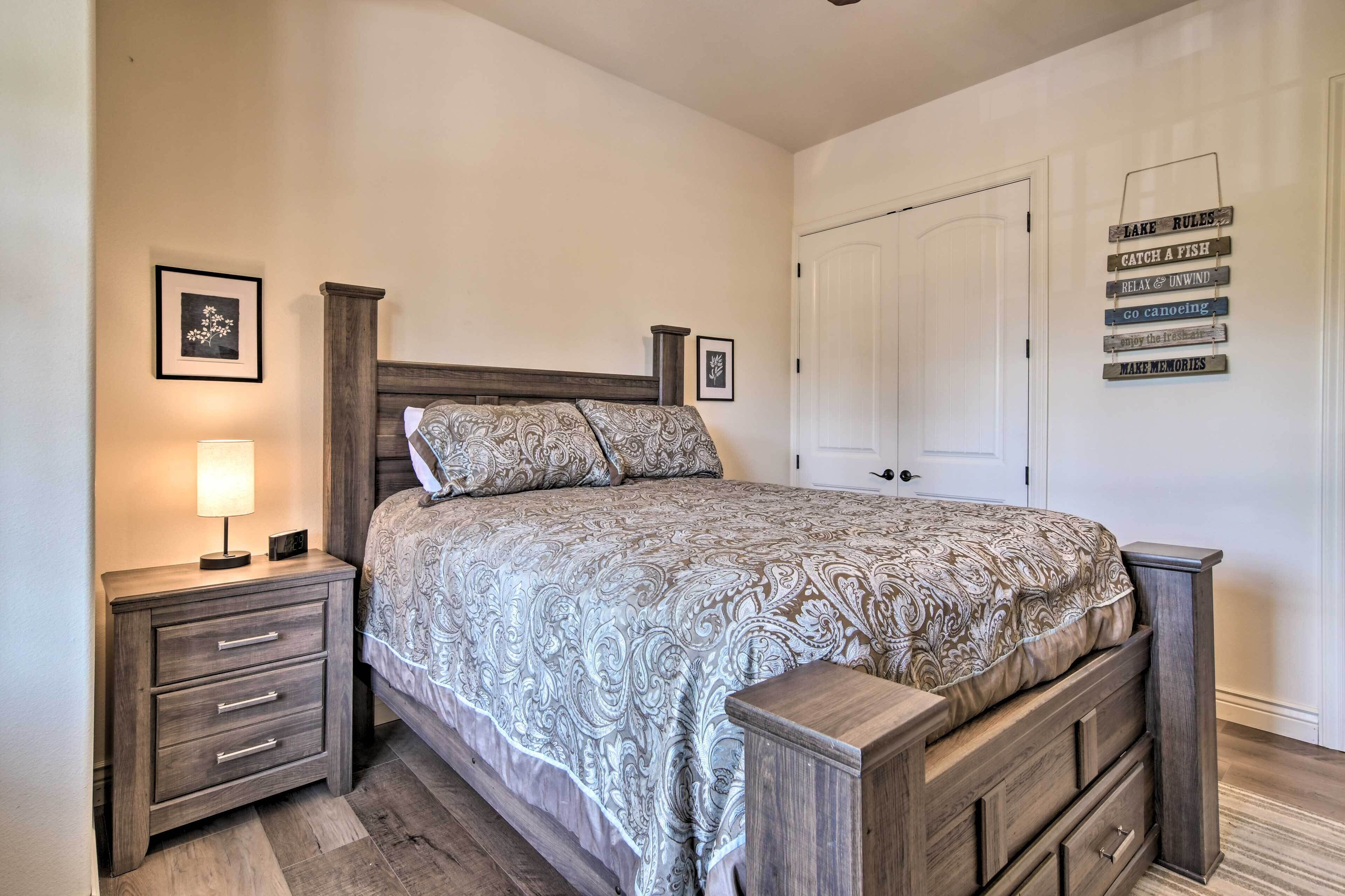 A bedroom with a wooden bed, a nightstand with drawers, a bedside lamp, and a wall with framed pictures and a sign.