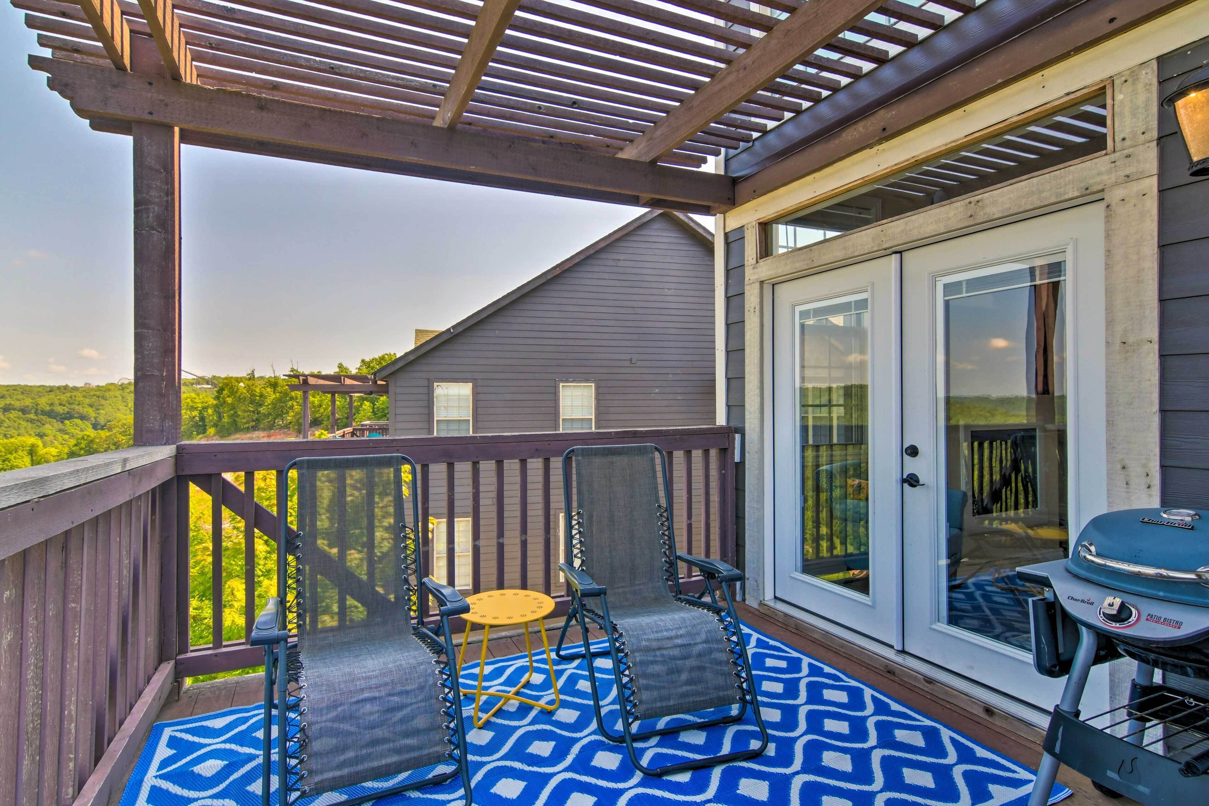 A covered deck with two chairs and a table, a barbecue grill, and a view of a house and greenery.