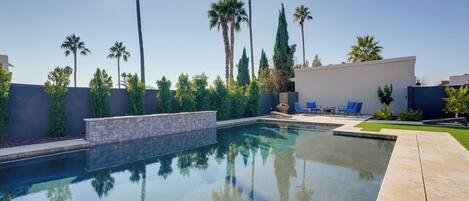 A modern outdoor pool area with clear blue water, surrounded by palm trees and a stone wall.