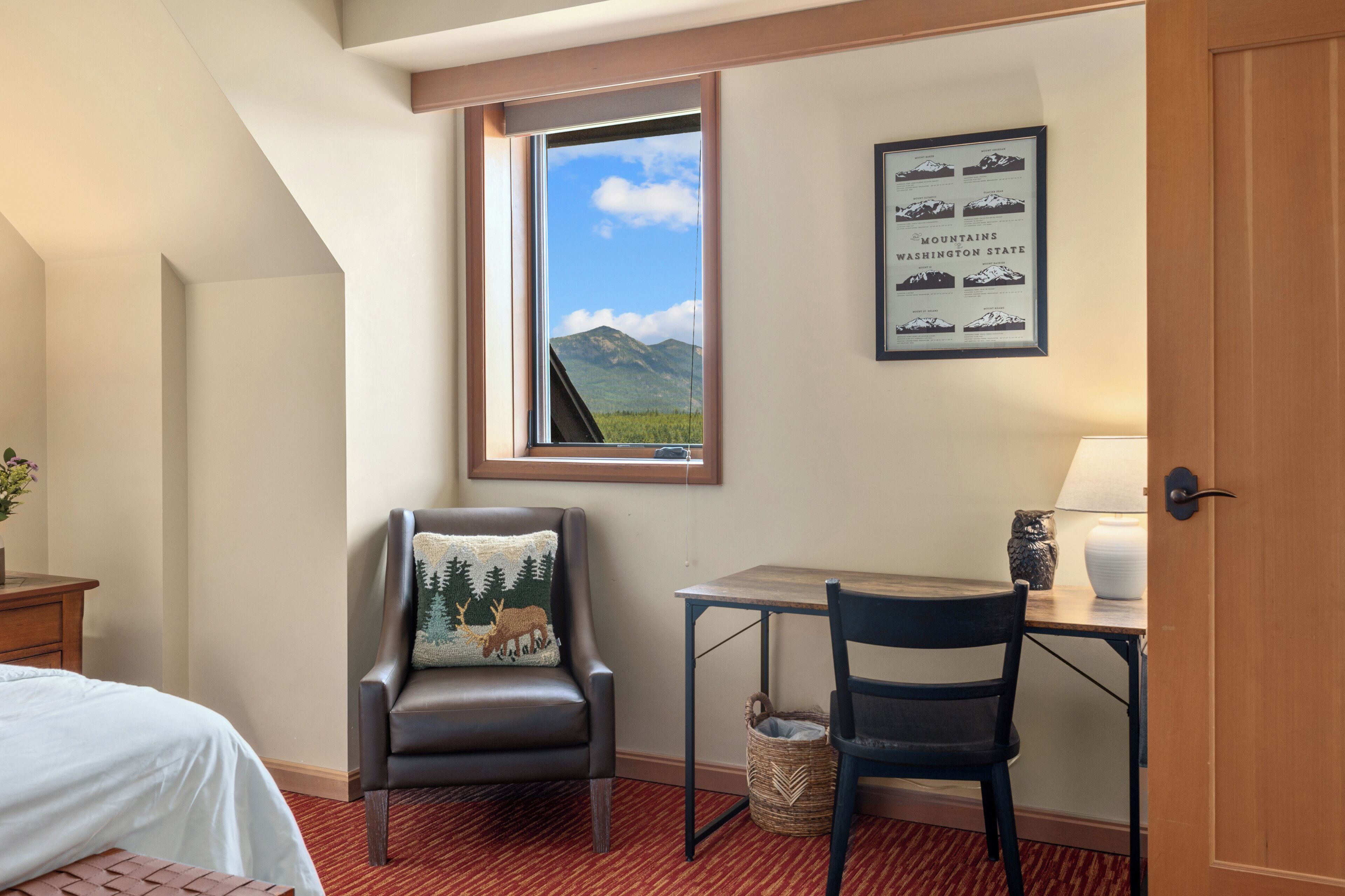 A hotel room with a bed, a desk, a chair, a window with a mountain view, and a framed picture of mountains.