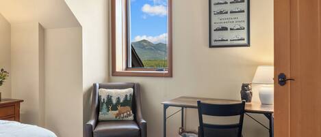 A hotel room with a bed, a desk, a chair, a window with a mountain view, and a framed picture of mountains.
